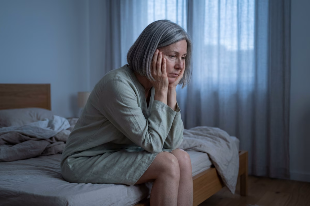 Femme de 53 ans assise au bord du lit à l'aube, épuisée par une nuit d'insomnie liée à la ménopause